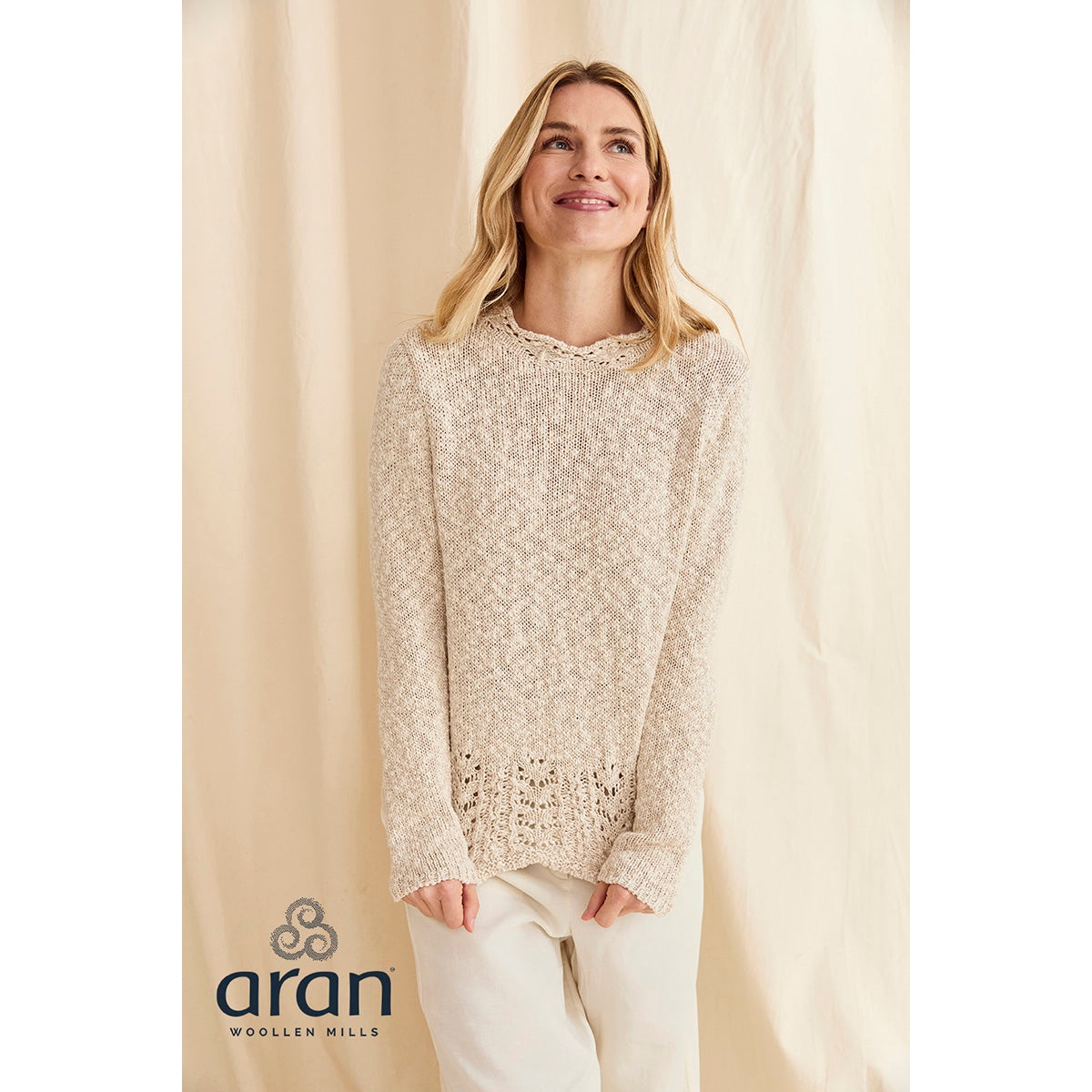Smiling woman wearing a cream knit sweater with a lace hem and white pants against a beige backdrop.