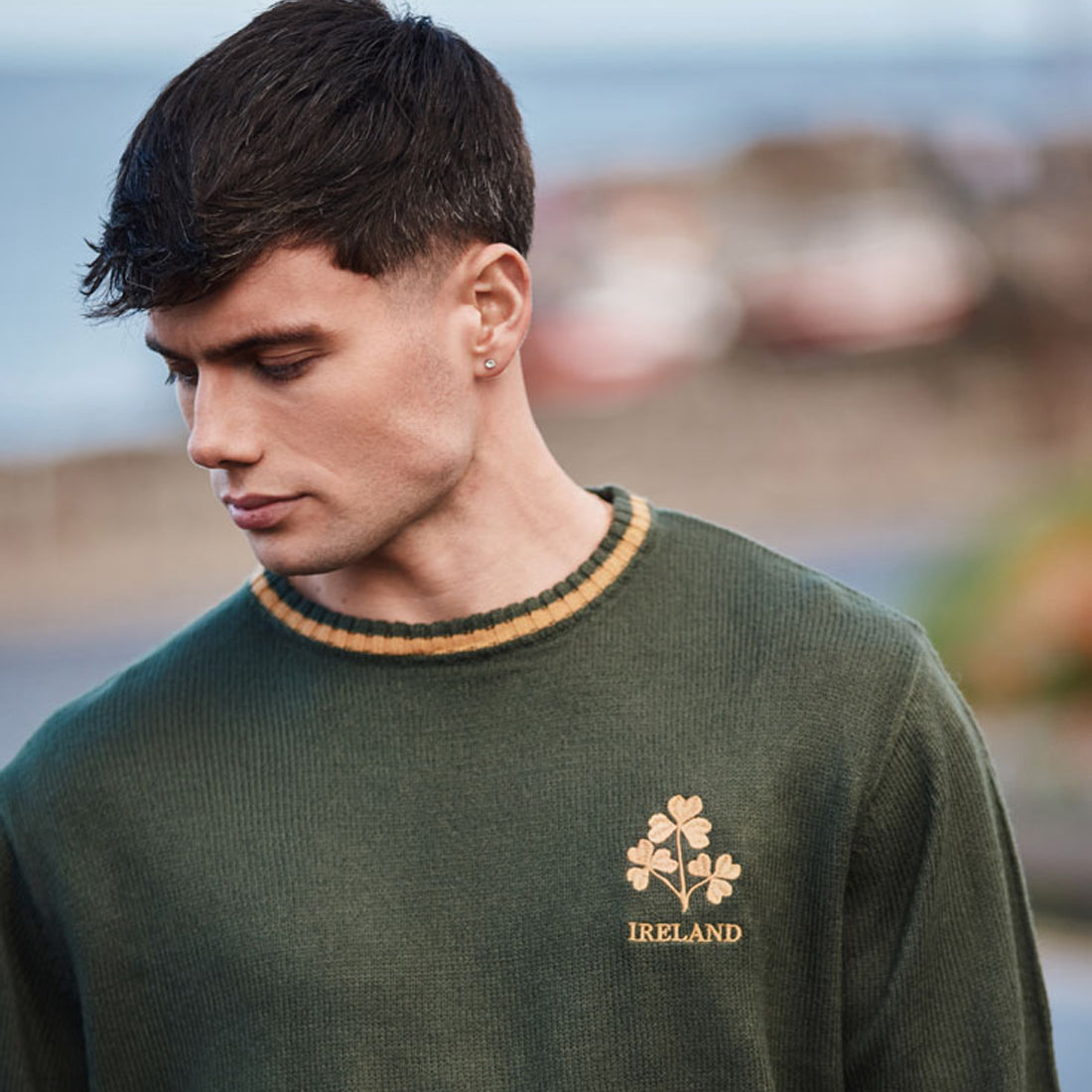 Man wearing bottle green crew neck jumper with orange striped collar and gold shamrock crest marked IRELAND text