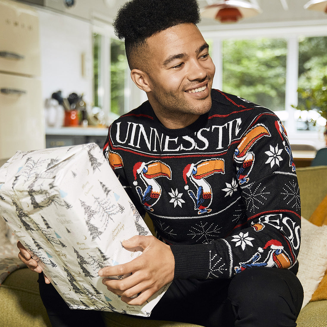 man with black hair sitting on a green couch and looking to the left in a room holding a present wrapped in white and silver wrapping paper wearing a black sweater with toucans wearing red Christmas hats and Guinness time written in white