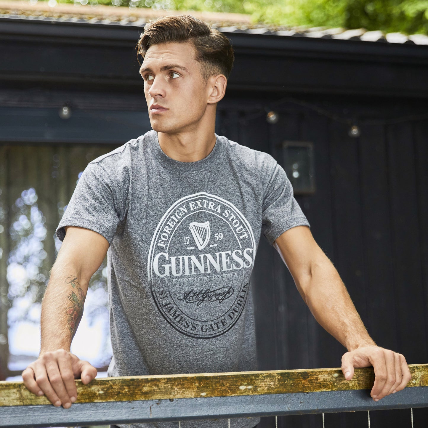 Young man wearing gray Guinness t-shirt leaning on railing outdoors