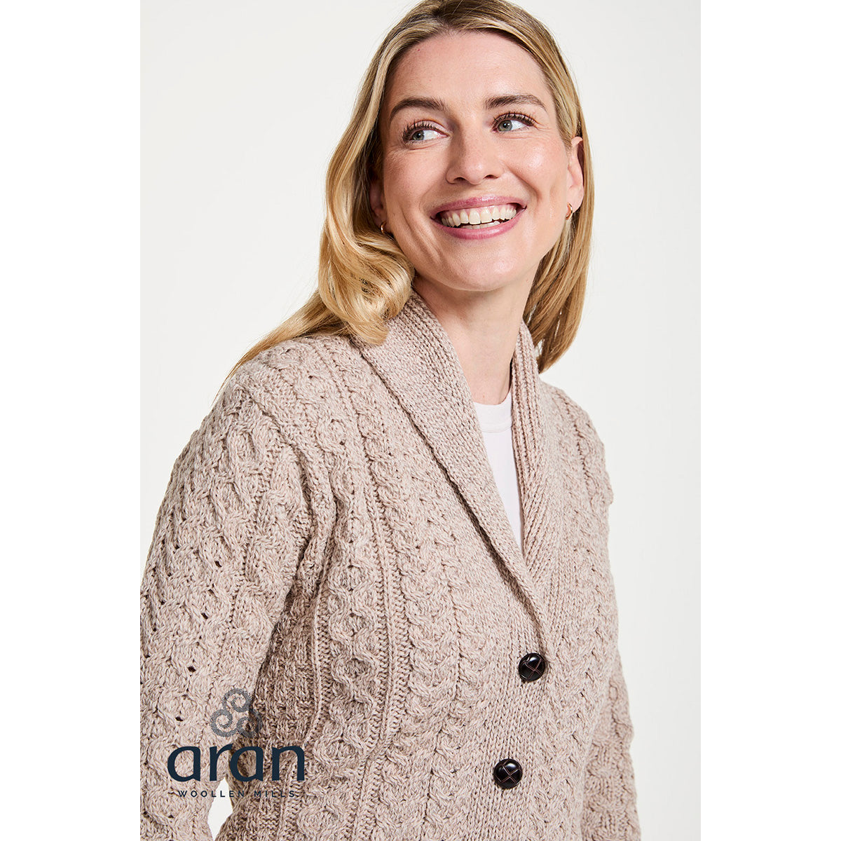Smiling woman wearing a beige shawl collar Merino Aran cardigan with chunky cable knit and dark buttons.