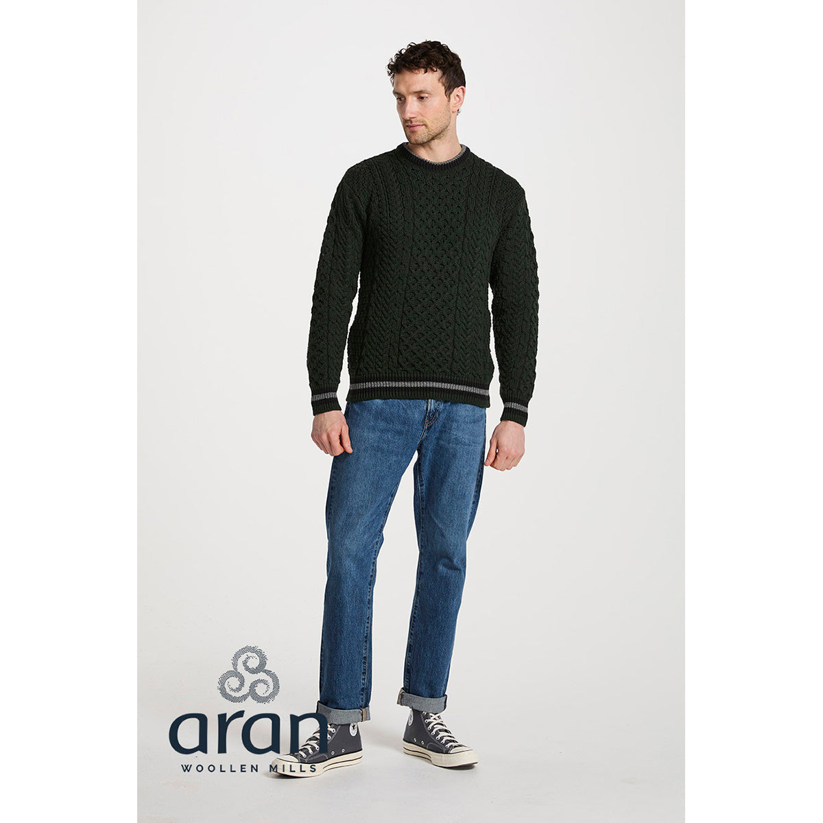 Full body shot of model in dark green Aran crewneck sweater with striped cuffs and hem, paired with jeans.
