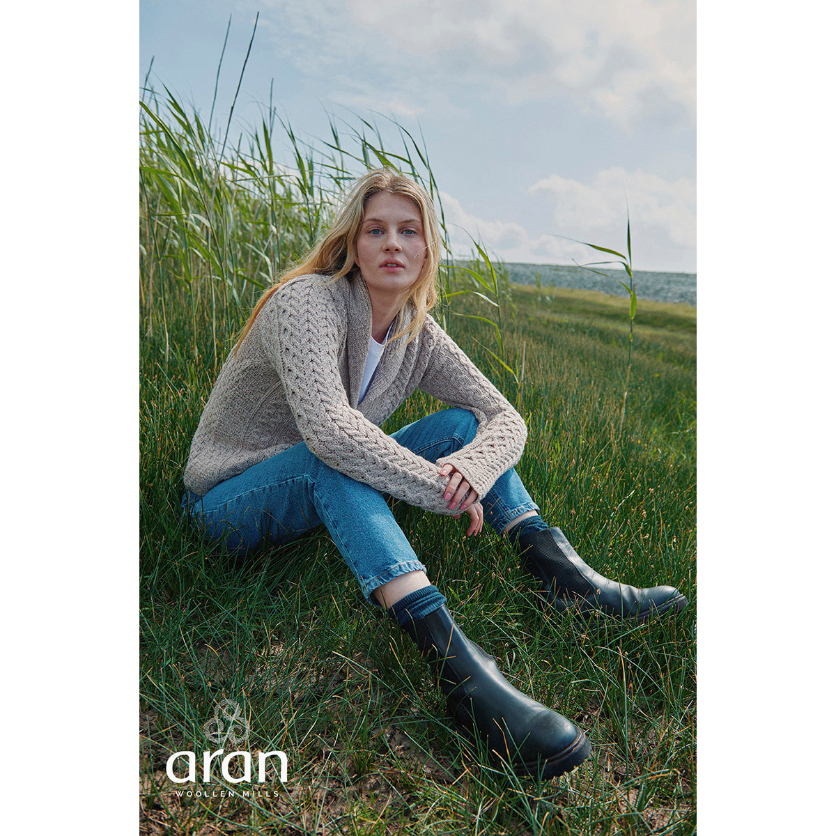 Model seated in tall grass wearing beige Tourmakeady shawl collar Merino Aran cardigan with jeans and boots