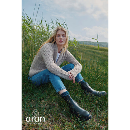 Model seated in tall grass wearing beige Tourmakeady shawl collar Merino Aran cardigan with jeans and boots