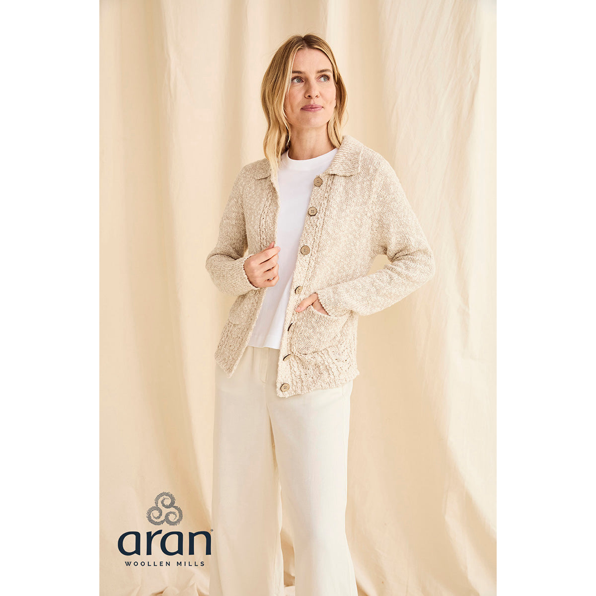 Full-length view of cream cardigan on a model against a beige backdrop with the brand logo at the bottom left