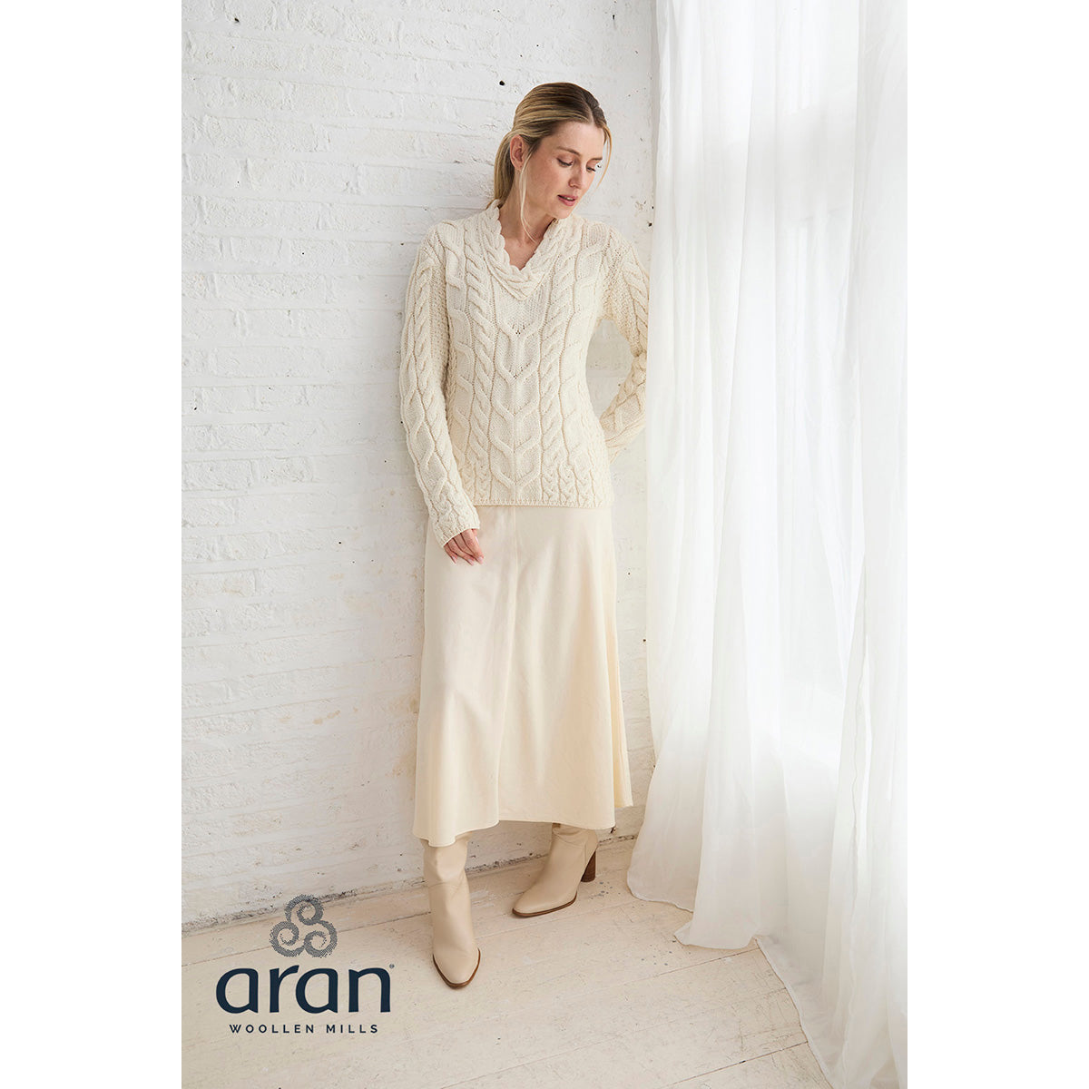 Model wearing cream Tralee Merino sweater with a matching skirt and knee high boots by a window