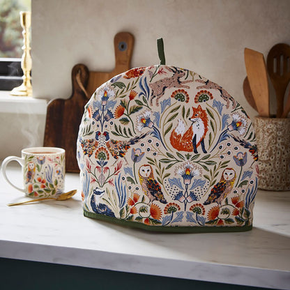 Tea cosy with a vibrant woodland print featuring foxes and owls on a kitchen counter beside a mug