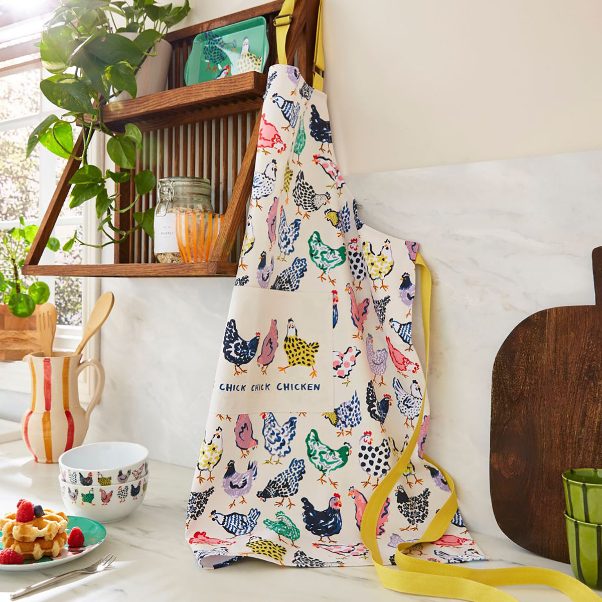 Cotton apron with colorful chicken prints and yellow trim hanging on a wooden rack in a bright kitchen