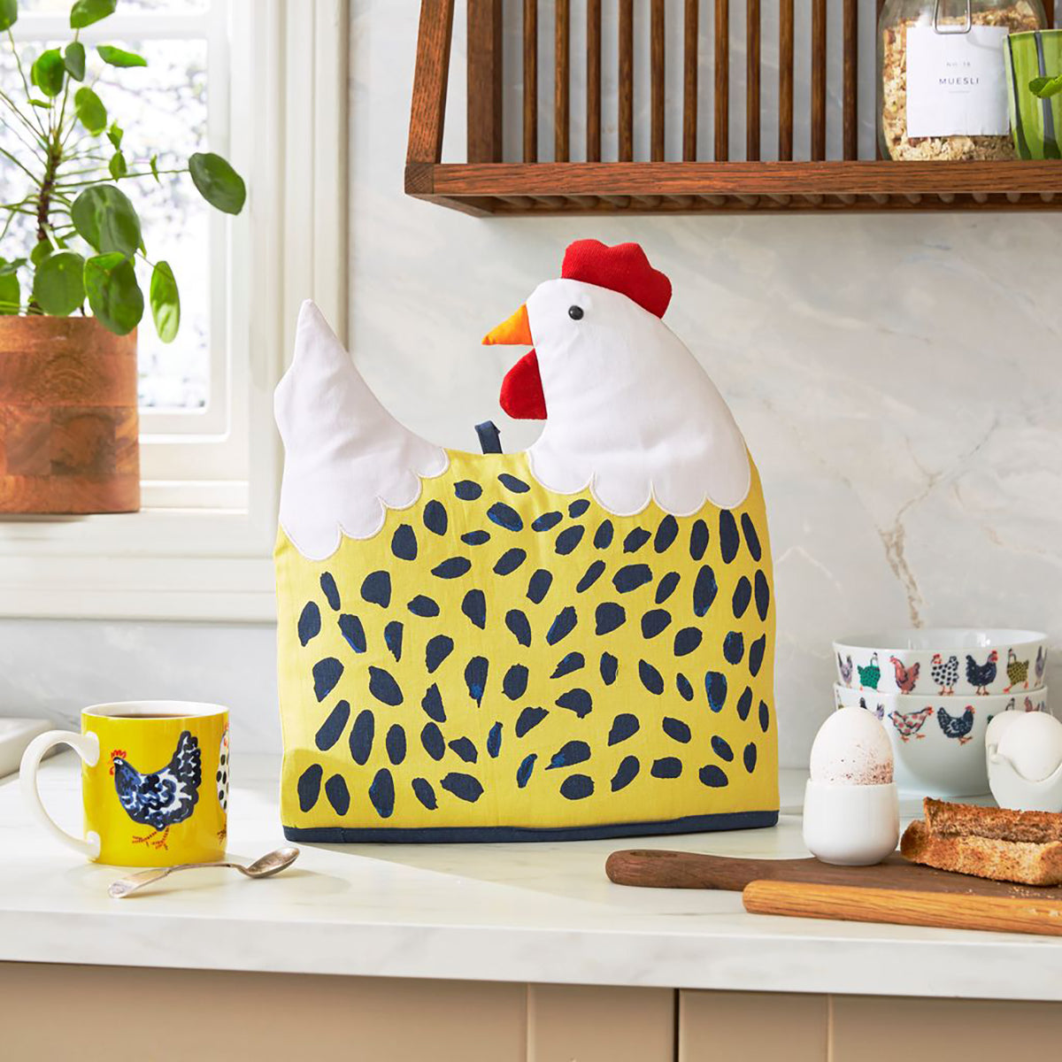 Chick chicken shaped tea cosy on a kitchen counter beside a yellow patterned mug and breakfast items
