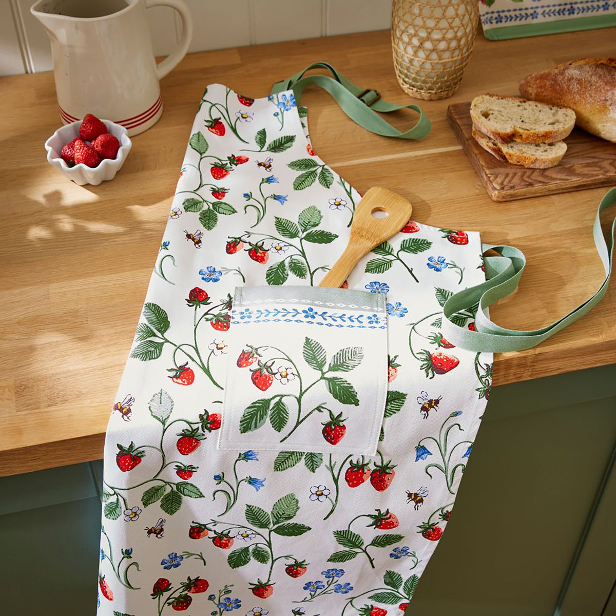 Strawberry Patch cotton apron spread on a wooden kitchen counter with a wooden spatula and bread nearby