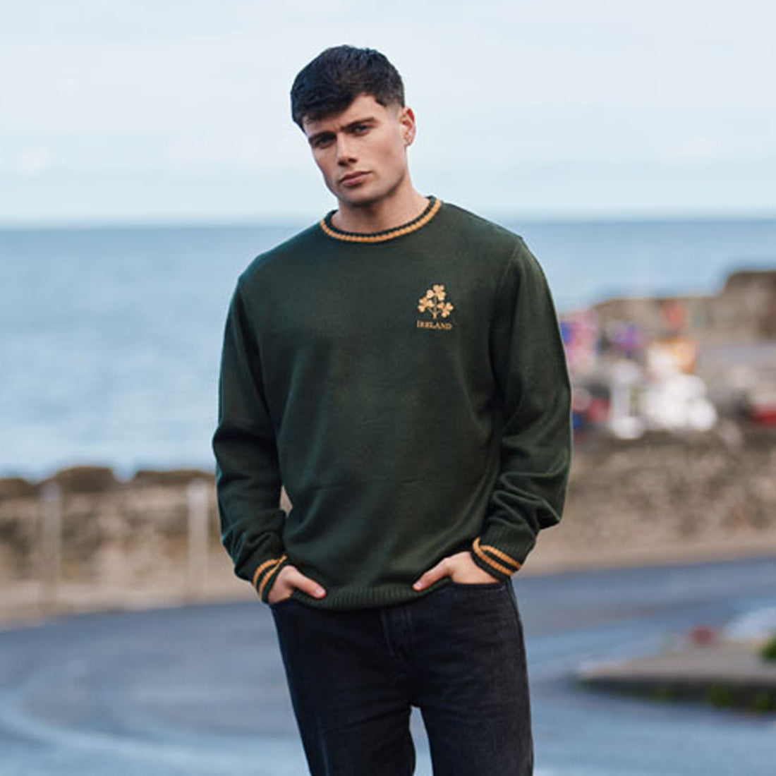 Male model wearing Ireland Bottle Green Three Shamrock Crew Neck Jumper by the coast