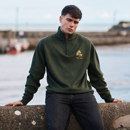 Male model in bottle green three shamrock jumper with quarter zip, seated on a seawall by the water.