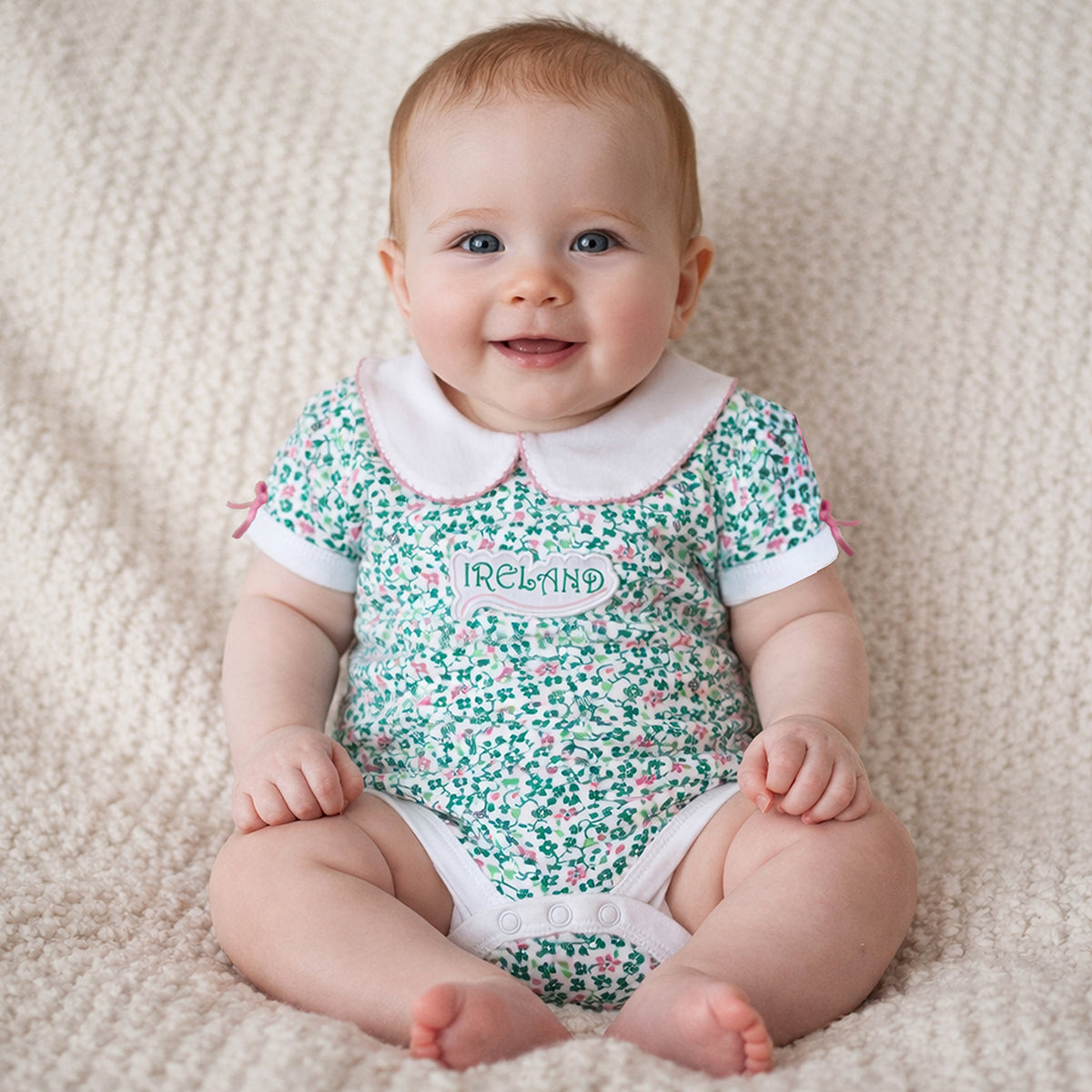 Smiling baby wearing a shamrock print vest with a white collar on a beige textured blanket