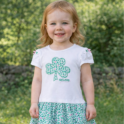 Smiling toddler wearing a white shamrock applique dress with a floral print skirt outdoors