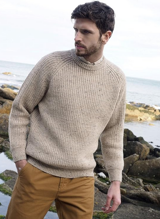 Man wearing beige knit sweater and brown pants standing near rocky shoreline