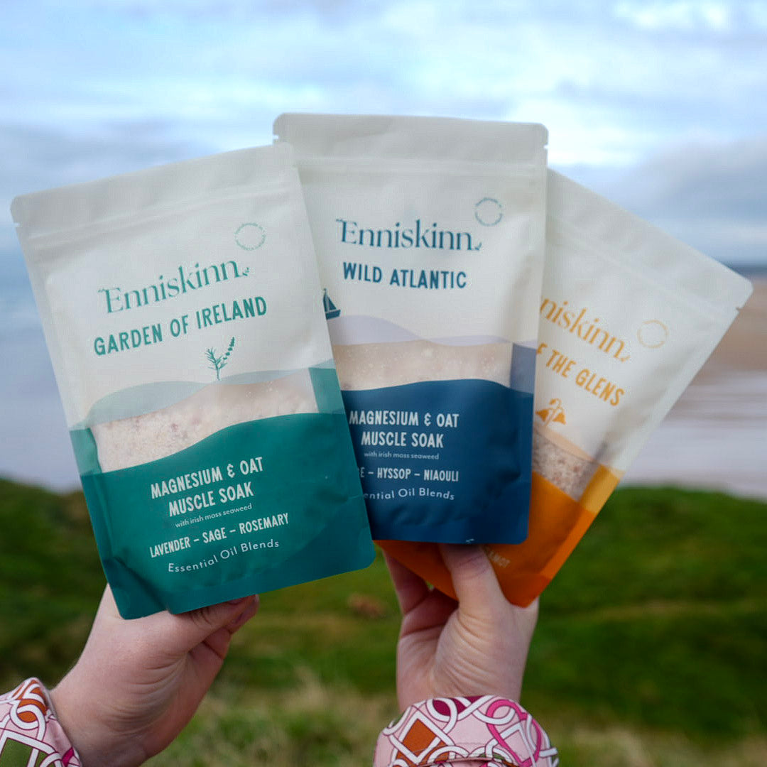 Three Enniskinn Garden of Ireland Magnesium and Oat Muscle Soak pouches held by hands outdoors near a coastal landscape