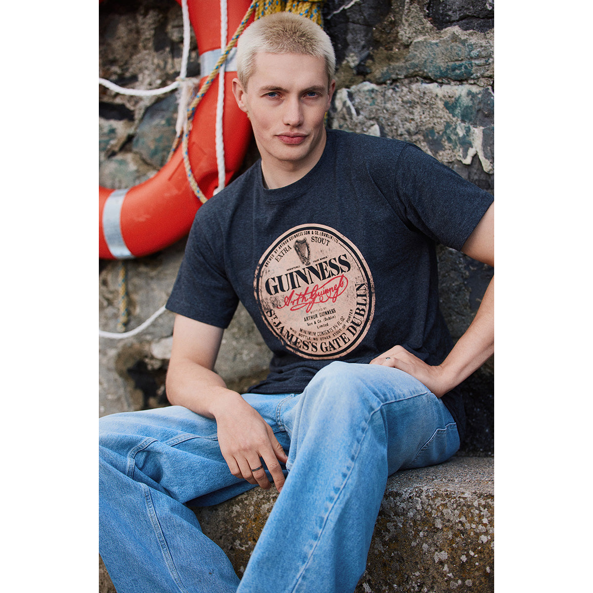 Male model in a charcoal Guinness T shirt with a circular vintage logo poses beside a rocky wall
