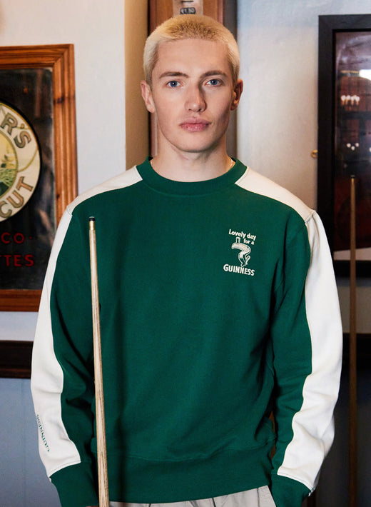 Young man wearing a green Guinness Toucan sweatshirt with white sleeves indoors beside vintage decor.