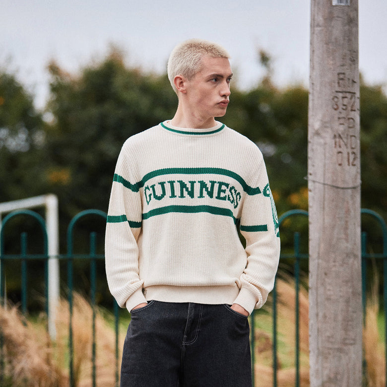 Cream Guinness crew neck knit jumper with green stripes and GUINNESS lettering worn by a man outdoors.