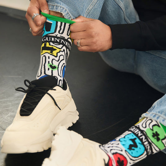 Person wearing Guinness Multi Doodle Socks with sneakers, adjusting the cuff.