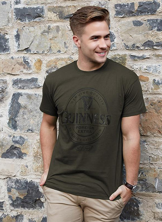 Young man smiling wearing dark green Guinness t-shirt standing by stone wall