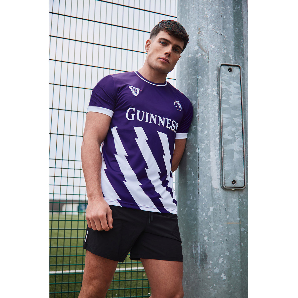 Male model wearing a purple Guinness Premier League football top with white trim, leaning against a metal pole beside a fence.