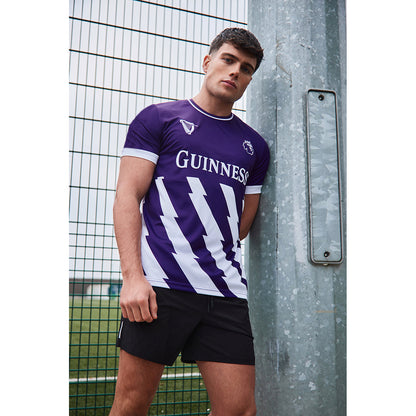 Male model wearing a purple Guinness Premier League football top with white trim, leaning against a metal pole beside a fence.