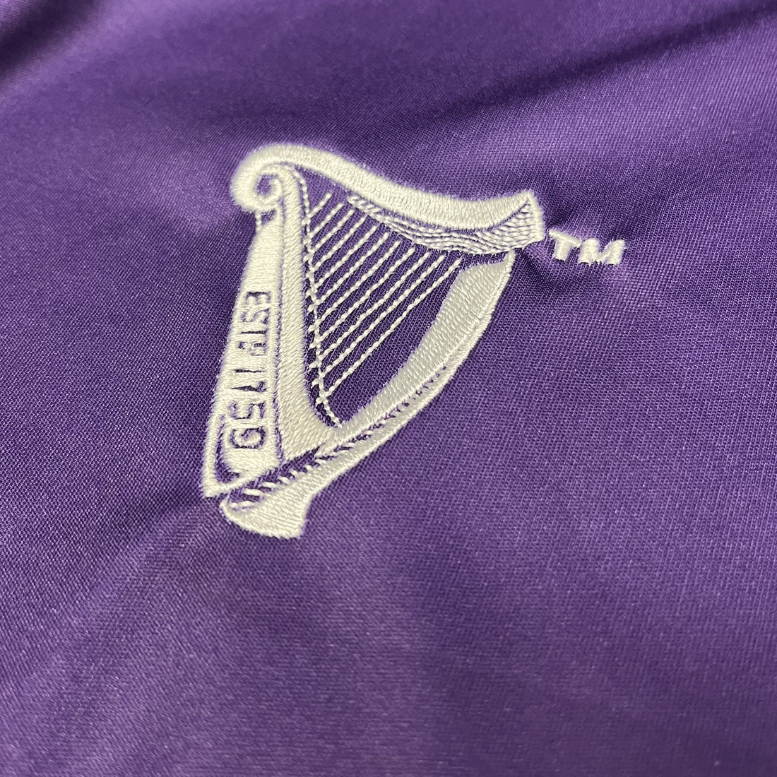 White embroidered Guinness harp crest on purple fabric of the football top.