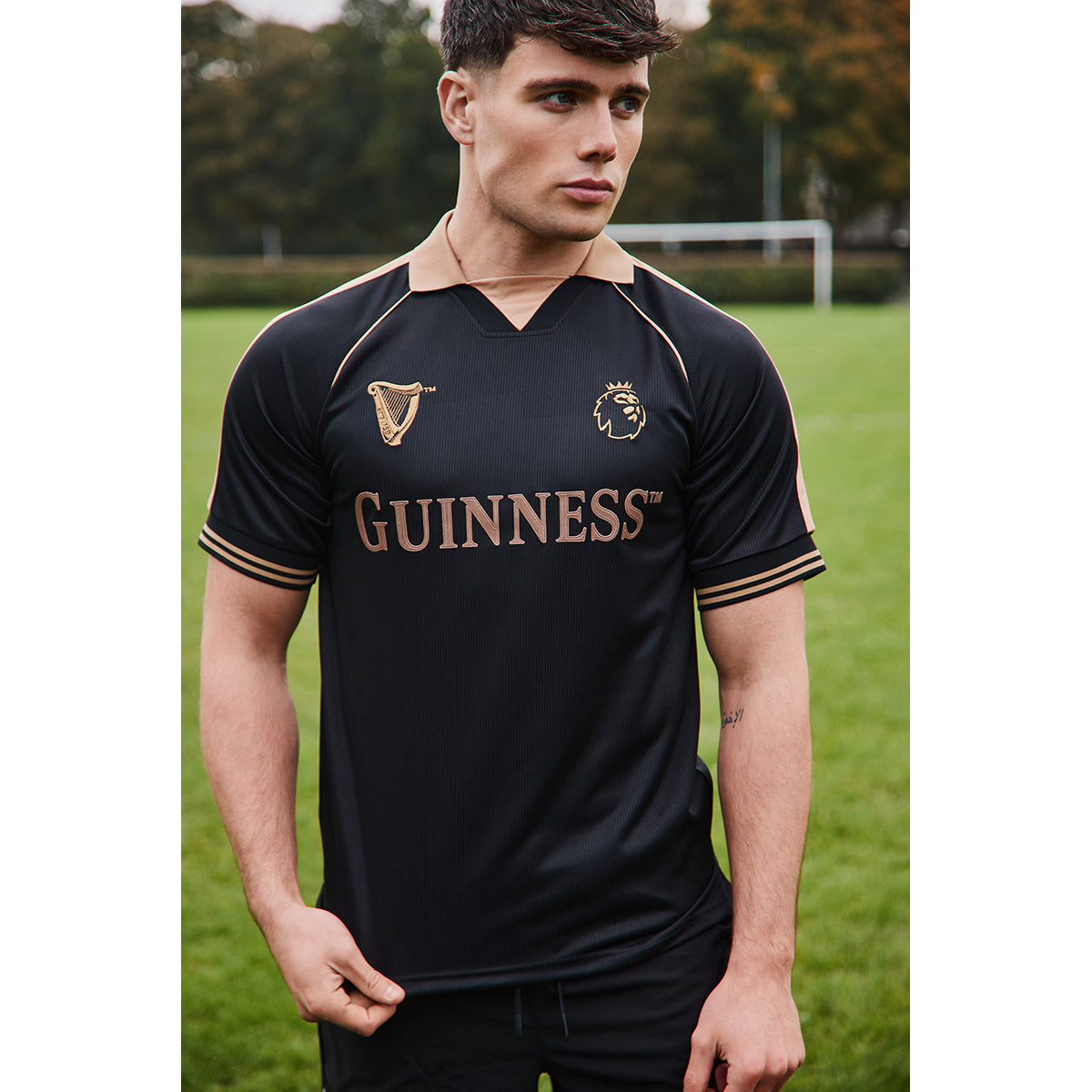 Guinness black football top with copper trim and gold lettering worn by a male model on a grassy field