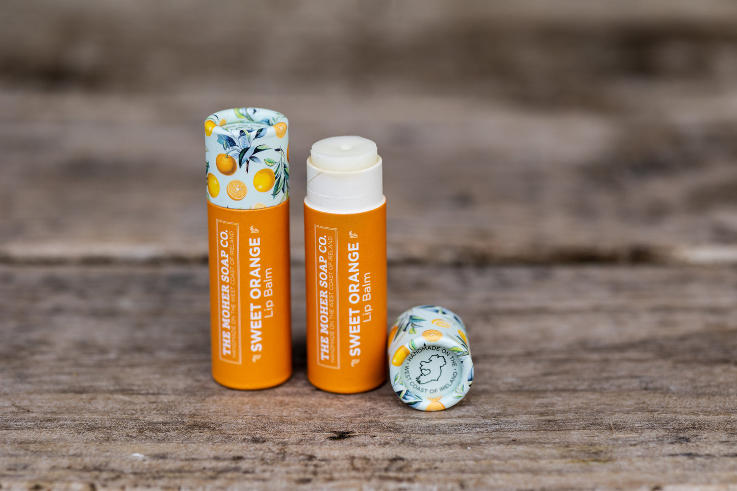 Sweet Orange Lip Balm set including two orange tubes and a citrus patterned cap on a wooden surface.