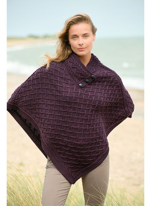Woman on a beach wearing a plum merino poncho with a chunky lattice knit and three front buttons.