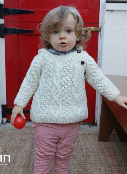 Toddler wearing cream cable knit sweater holding red ball indoors