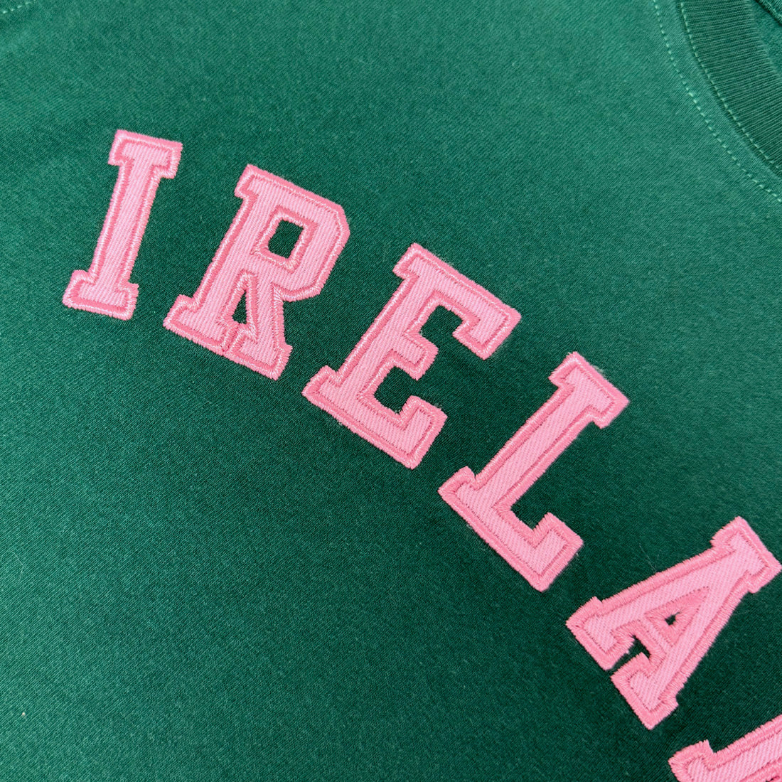 Close-up of pink embroidered Ireland lettering on green T-shirt fabric.