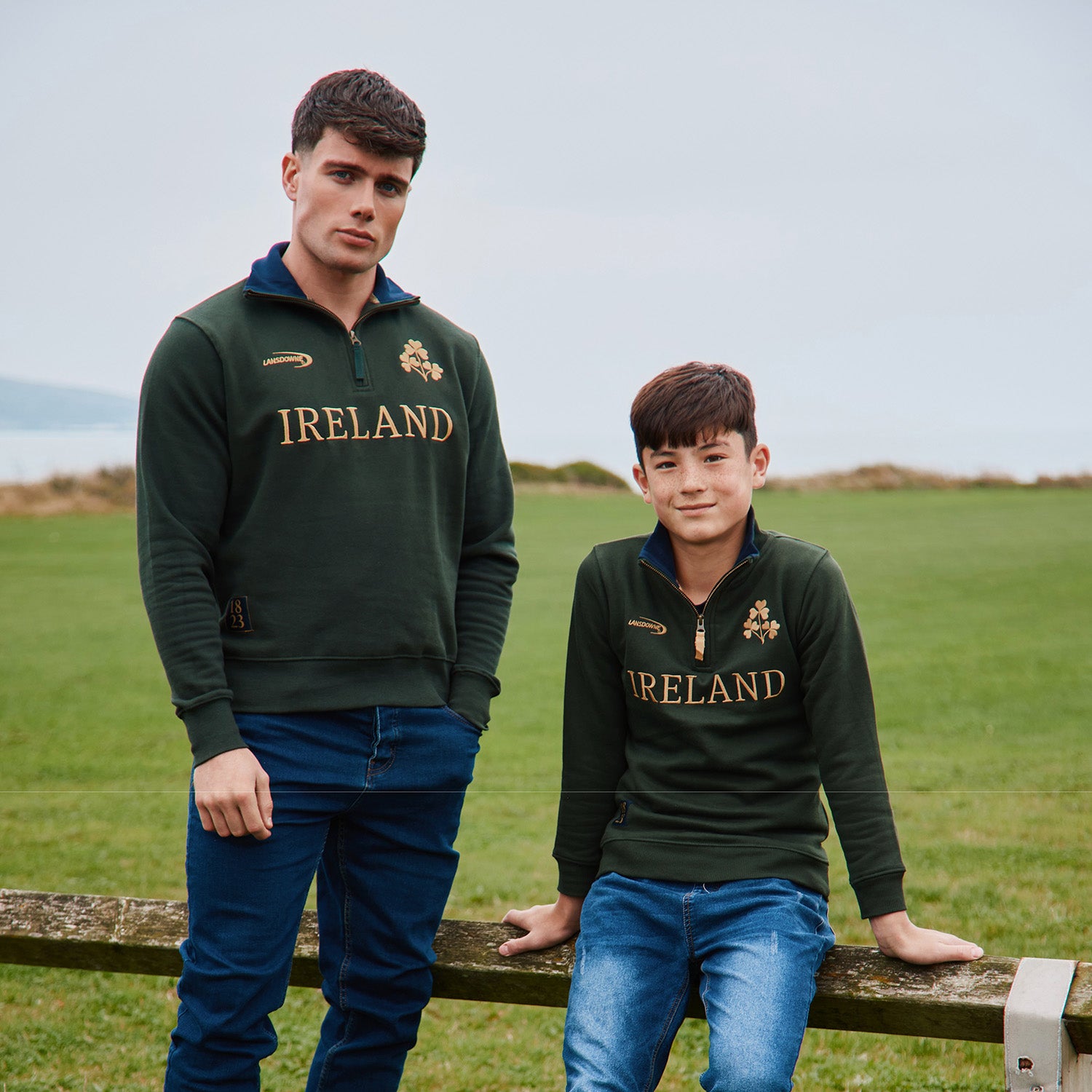 Two men in Lansdowne Ireland bottle green and gold fleece tops with crest posing outdoors in a field.