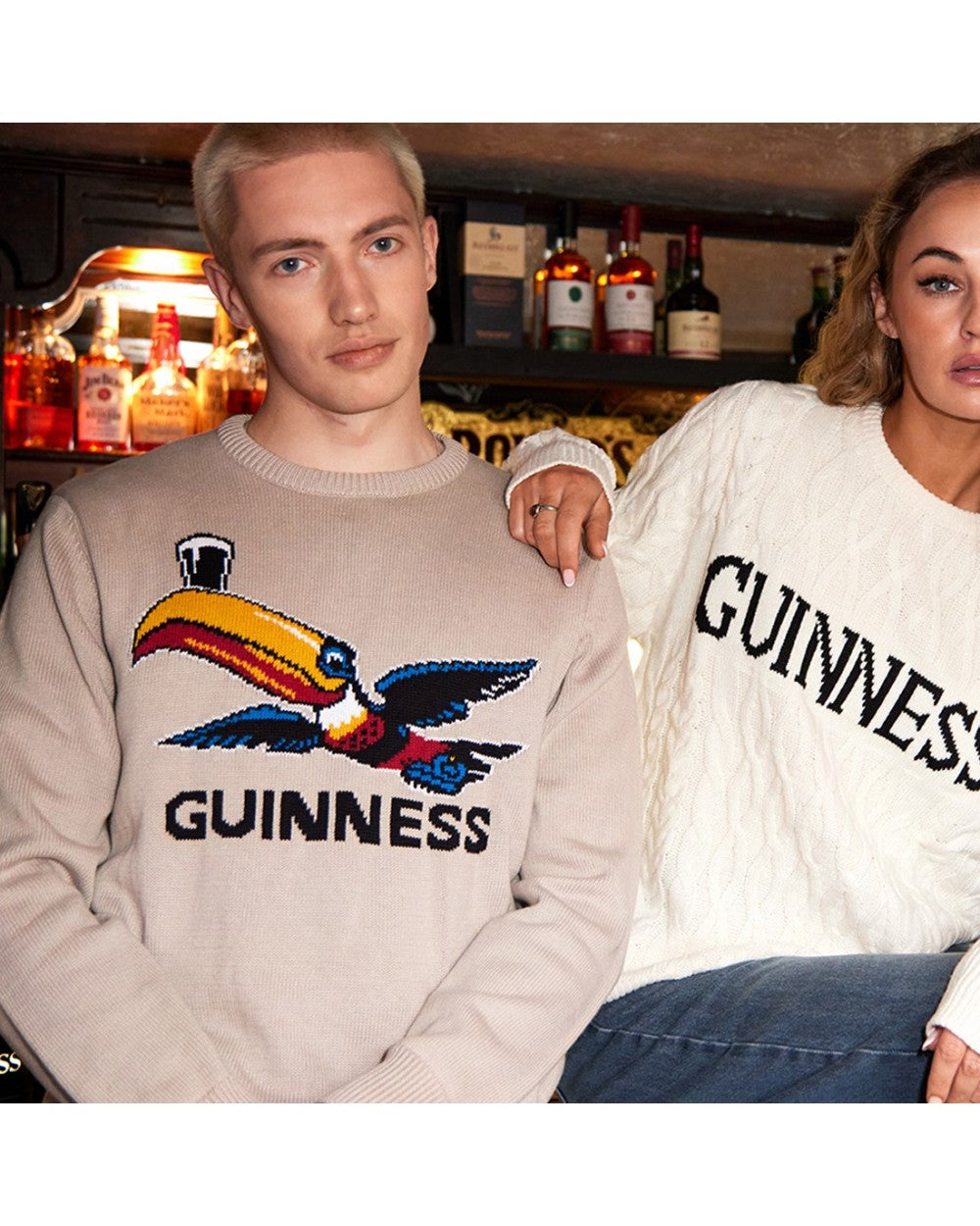 Couple wearing Guinness jumpers in a bar setting; man in beige toucan sweater and woman in white knit with Guinness lettering