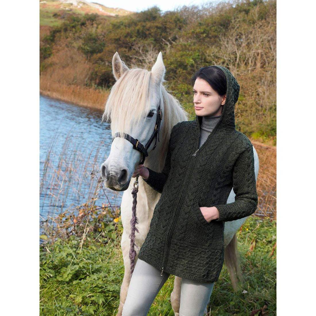 Woman in dark hooded sweater holding white horse near a river